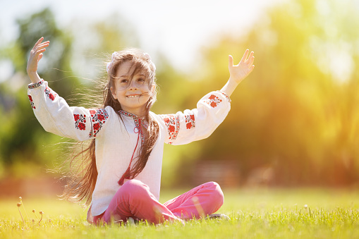 Cute smiling girl in the Ukrainian national wear on the green meadow. Portrait of fun happy girl with outstretched hands, sitting on the green grass.