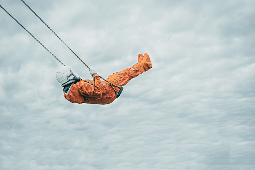 Astronaut wearing orange space suit and space helmet on a swing against cloudy sky. Cosmonaut flying on a swing outdoors