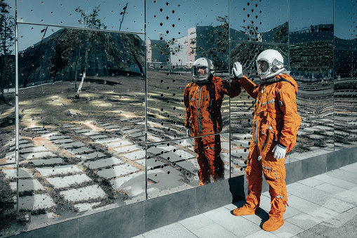 Astronaut wearing orange spacesuit and space helmet near a huge mirror wall of building outdoors