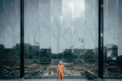 Astronaut wearing orange spacesuit and space helmet standing near the building of a space center outdoors. Cosmonaut in orange space suit posing agains a huge mirror wall outdoors