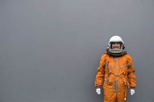 Astronaut wearing orange space suit and helmet standing against gray wall background. Happy cosmonaut on a grey background.