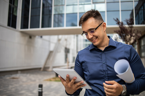 A young businessman walks in a park around a business complex and carries his digital tablet and business projects