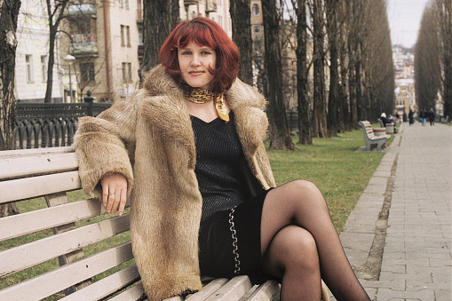 Young beautiful woman in a fur coat sits on a park bench. File source is film.