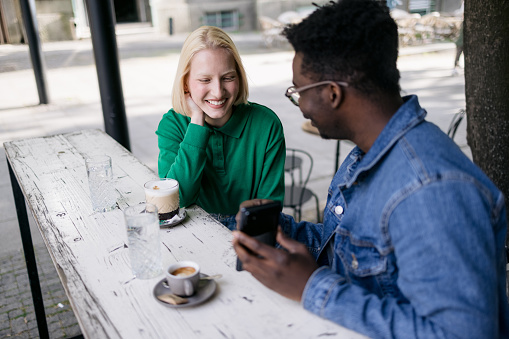 A diverse pair of happy friends meeting in an outdoor coffee shop, talking and catching up, young man showing his female friend something on his smart phone