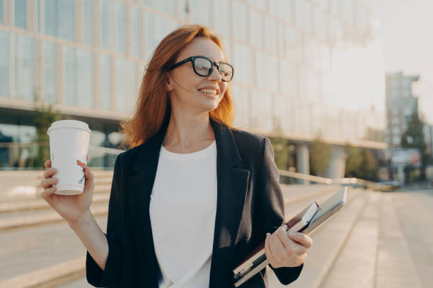 istock Overjoyed happy ginger woman in spectacles drinking coffee and holding laptop while walking outdoors 1397962055