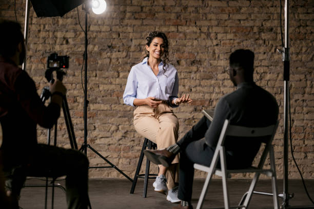 Giving an interview in a modest studio Happy attractive woman sitting on a bar stool and giving an interview to a male journalist in front of her, gesturing with her hands while she's talking and smiling stage set stock pictures, royalty-free photos & images