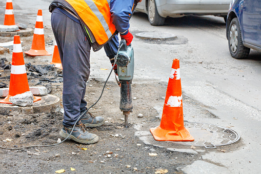 A road worker works with an electric jackhammer on a road section fenced with traffic cones.
