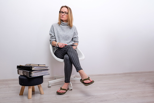 blond woman with laptop on her lap sitting next to a stool with document folders, remote work