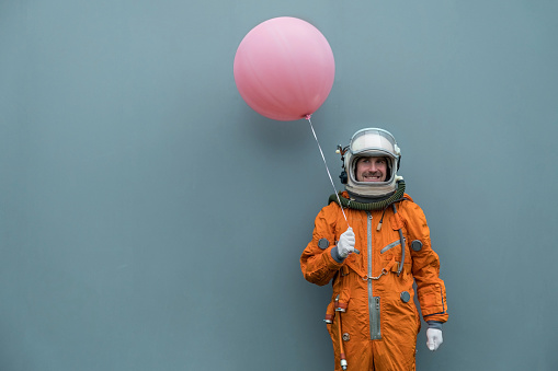 Astronaut wearing orange space suit and space helmet holding a pink inflatable balloon against gray wall background. Happy cosmonaut with a pink helium air balloon