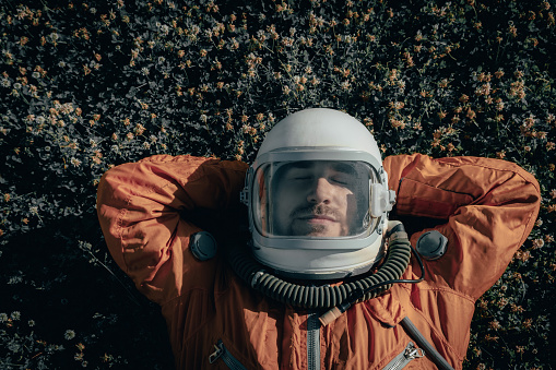 Closeup of cosmonaut wearing space suit and space helmet taking a nap while lying on green grass outdoors. Portrait of astronaut sleeping on meadow with flowers. View from above