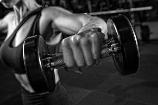This is a close up black and white photo of a woman working out in a dark moody lit gym  with dumbells