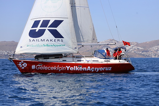 Bodrum,Mugla, Turkey.07 May 2022: sailor team driving sail boat in motion, sailboat wheeling with water splashes, mountains and seascape on background. Sailboats sail in windy weather in the blue waters of the Aegean Sea, on the shores of the famous holiday destination Bodrum. Men and women on a sailboat in a very cold weather continue to cruise. Sailing crew on sailboat during regatta. Sailing in the wind through the waves at the Sea. Close up of sailing boat, sail boat or yacht at sea with white sails.