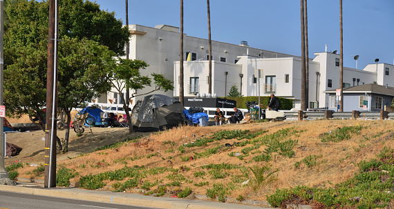 San Pedro, California, USA - April 29,2022 : A homeless campsite overlooking the Port of Los Angeles with three men having their morning coffees and very expensive residences in the background.