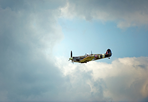 Supermarine Spitfire Mk VB fighter flying in vicinity of Duxford, Camdridgeshire, England. Solo classic wartime single seat fighter aircraft, best known for tis famous role as principal RAF fighter in the Battle of Britain, flying in moody sky cloudscape over the fields of England. This marque is one of the later wartime models with clipped wings for additional speed