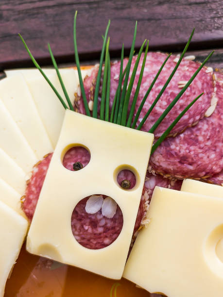 様々なチーズ、ソーセージ、肉のスライスが皿の上に並べられています。穴の開いたチーズのスライスを作る芸術は、髪の毛を持つ人間の顔のように見えます。 - cheese making ストックフォトと画像