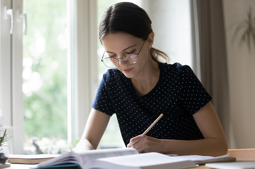 High school student girl in eyeglasses sit at table looks serious and focused learns subject, makes notes on copybook. Self-education process, develop skills, improve knowledge, do assignment concept