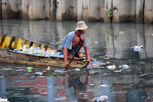 Jakarta, Java, Indonesia: canoe with man picking plastic bottles from the heavily polluted waters of Kali Besar canal, Kota - bottles will sold for recycling - circular economy opportunities.