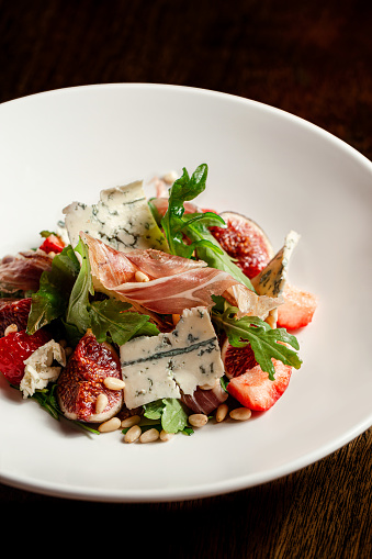 Salad with hamon ham, figs, blue cheese, arugula and strawberry on a plate. Dark Wooden background.