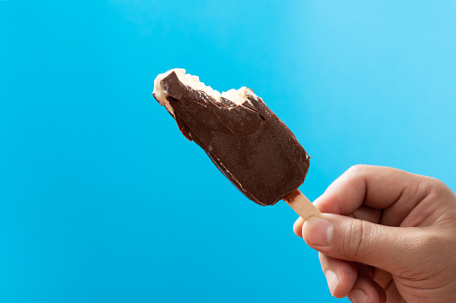 man holding a chocolate outer popsicle with couple of bites on blue background