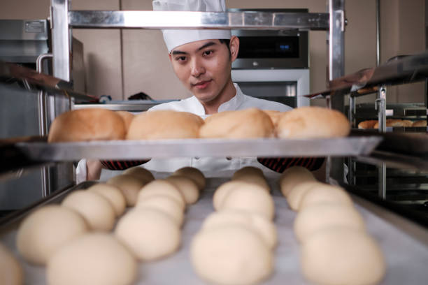 Young professional Asian male chef in white cook uniform with hat, gloves, and apron making bread from pastry dough, preparing fresh bakery food, baking in oven at restaurant stainless steel kitchen.
