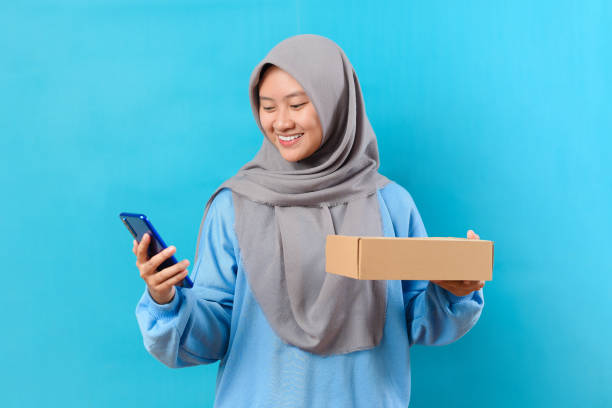 mulher de negócios millennial segurando caixa de pacotes para entrega e usando smartphone isolado em fundo azul - teenager smart phone young women teenagers only - fotografias e filmes do acervo