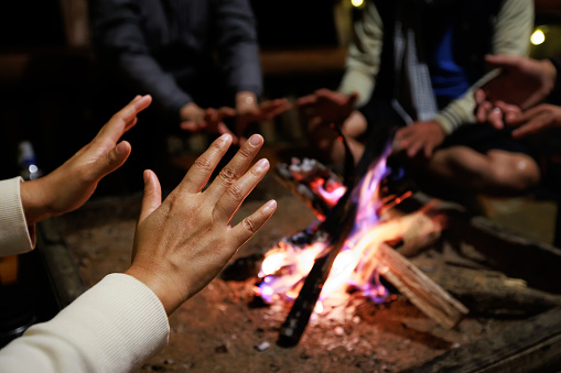 Close-up on hand of woman warming on campfire. Group of asian friends sitting around campfire in the night. Friends camping near bonfire relaxing on winter vacation at the night time.