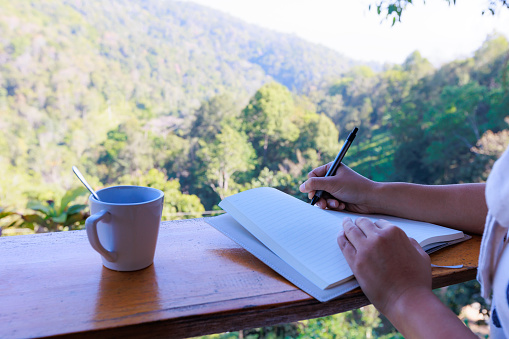 Asian woman sitting on balcony and writing idea on notebooks near fresh nature with hot drink in the morning time.