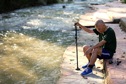 An Asian senior man is relaxing at nature reserve forest. He is seen wearing ankle weight for strength training.