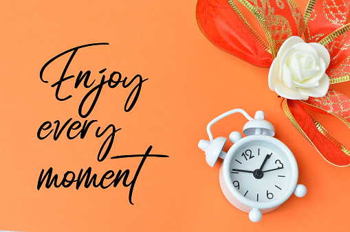 Clock and flower over orange background written with ENJOY EVERY MOMENT.