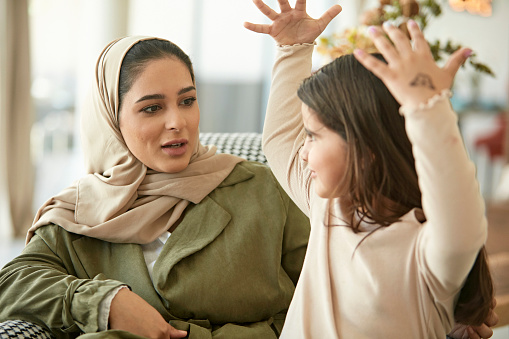 Waist-up view of late 20s woman in abaya and hijab smiling and talking with 6 year old daughter as they spend time together in family living room.