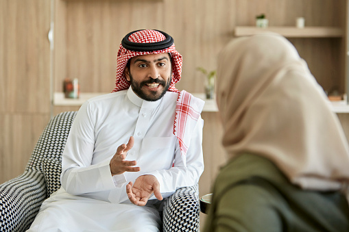 Over the shoulder view with focus on early 30s bearded man in traditional attire face to face and gesturing while talking with woman in living room.