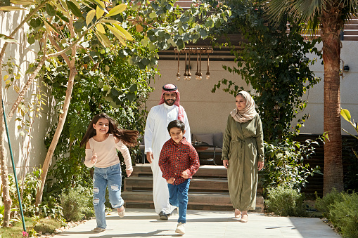 Full length front view of 6 and 7 year old siblings in casual clothing leaving shaded patio and running towards camera with smiling parents following behind.