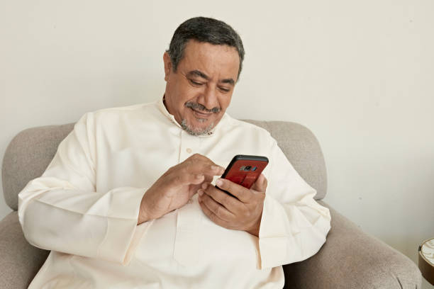 Mature Saudi man relaxing at home with smart phone Waist-up front view of contented Middle Eastern man wearing dish dash and sitting in living room armchair using portable device. arabian peninsula stock pictures, royalty-free photos & images