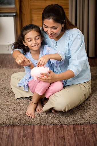 Happy mother and daughter inserting coin into piggy bank at home