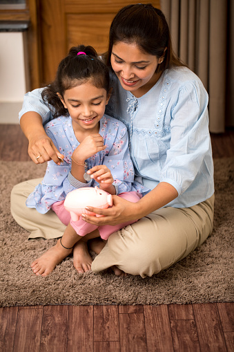 Happy mother and daughter inserting coin into piggy bank at home