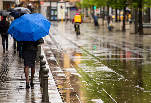 Unrecognizable woman with blue umbrella walking down city street in a rainy weather