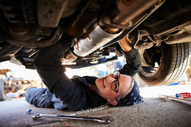 Smiling female mechanic working on the underside of a car Portrait of a young female mechanic smiling while working on the underside of a car up on a jack outside of her auto workshop mechanic working under car stock pictures, royalty-free photos & images