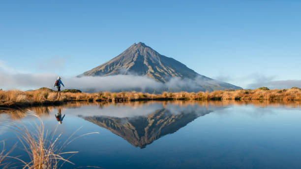 Woman hiking Pouakai circuit. Mt Taranaki reflected in the Pouakai tarn in morning sun. New Zealand. Woman hiking Pouakai circuit. Mt Taranaki reflected in the Pouakai tarn in morning sun. New Zealand. dormant volcano stock pictures, royalty-free photos & images