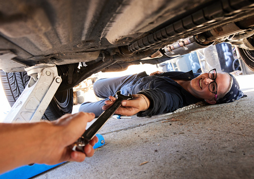 Smiling young female mechanic taking a wrench from someone while working underneath a car outside of her workshop