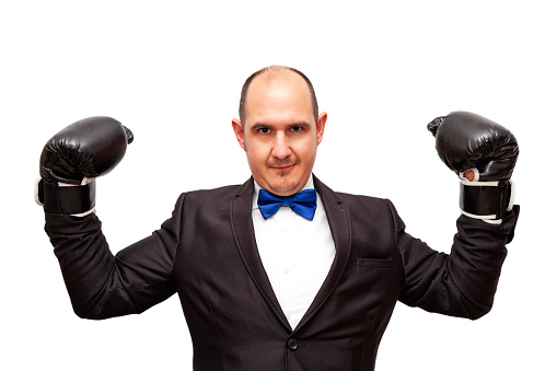 A bald Caucasian adult male dressed in a suit, shirt and bow tie and wearing boxing gloves is making a power gesture with his arms. The background is white.