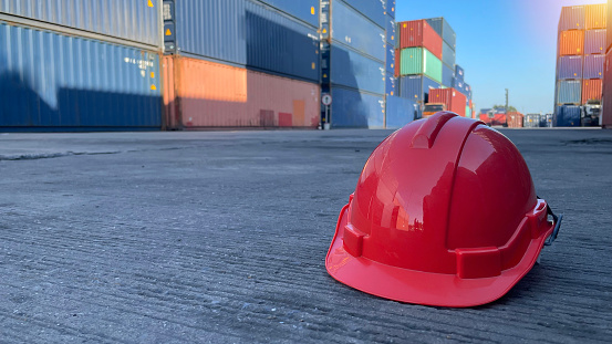 Red hard safety helmet hat for engineer or worker on concrete ground with container blurred image background (safety concept)