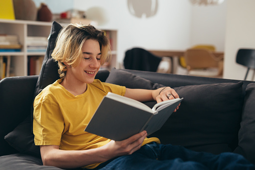 young woman is reading a red book on the sofa. a girl in a yellow sweater and blue jeans