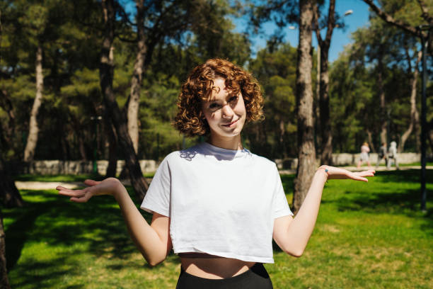 Unsure woman wearing white tees and black yoga pants shrugging shoulders over at outdoor. Attractive redhead woman looking to camera. Hesitating woman. Helpless feelings. Unsure look stock photo