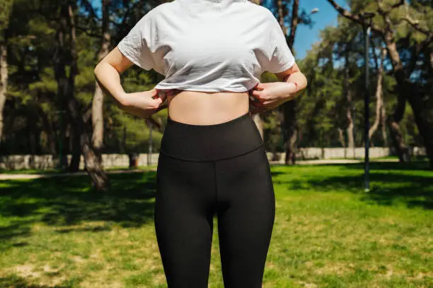 Photo of Young sportive woman wearing black yoga pant and white tees standing on a park bench.