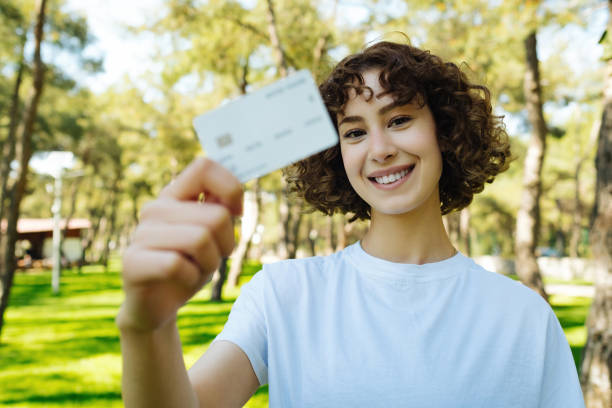 Redhead young smiling woman in white tee holding credit bank card or debit card on green city park background. Outdoors, sportive and online shopping concepts. Selective focus on woman. stock photo