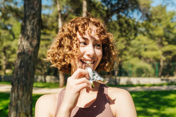 Closeup face of young sporty redhead woman wearing sport bra resting while biting a nutritive bar. Athletic woman eating a protein bar. Fitness beautiful woman eating a energy snack outdoor. stock photo