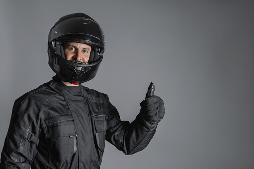 Safe ride concept. Confident Man wearing protective black motorcycling equipment, with thumb up, indoors. Studio shot, grey background. Copy-space for insurance advertisement.