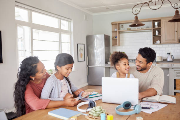 mixed race family sitting together doing homework and using digital devices at kitchen table. couple sitting at home with two daughters and using modern technology for online learning - couple laptop computer digital tablet imagens e fotografias de stock