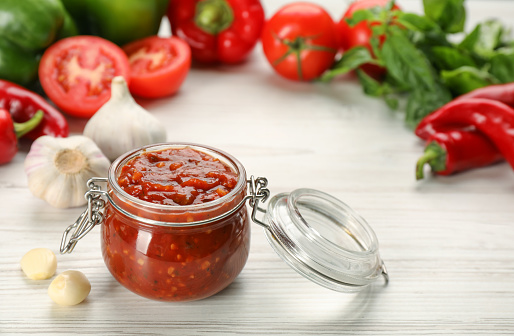 Glass jar of tasty adjika and ingredients on white wooden table, space for text