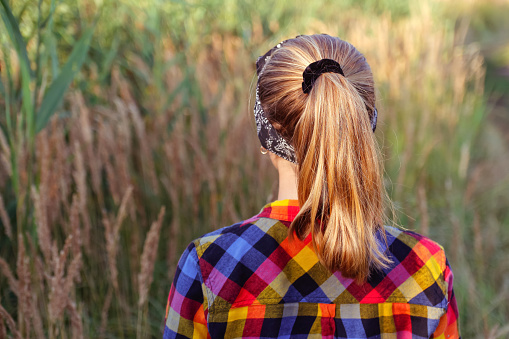 Defocus ponytail hairstyle. Teen or preteen girl walking on nature background and standing back. Little kid girl. Green meadow. Generation z. Autumn, summer. Bandana. Lifestyle. Sunny. Out of focus.